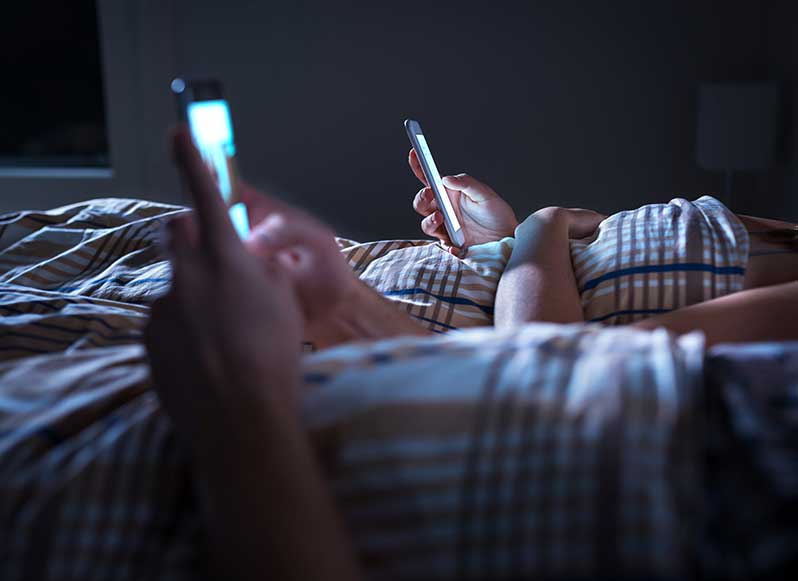Bored distant couple ignoring each other lying in bed at night while using mobile phones.